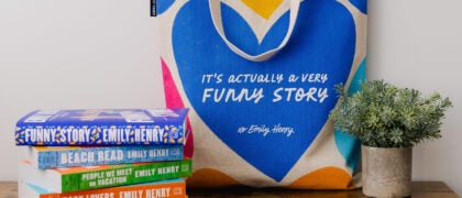 The image shows a colorful tote bag with the text "It's actually a very funny story by Emily Henry" alongside a small potted plant. Next to the bag is a stack of books by Emily Henry, with titles including "Beach Read" and "People We Meet on Vacation.